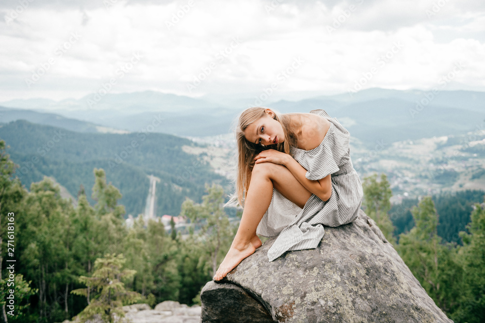 Naklejka premium Lonely young girl sits at stone on top of mountain with far view at hills and cloudy sky and hugs her legs with head lying on knees. Beautiful blonde woman outdoor summer portrait. Female at nature.