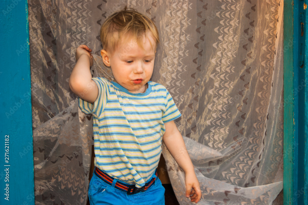 In early spring, in the village, a small blond boy of three years old, chumazenky, comes out of his village house, whose door is covered with insect tulle In early spring, in the village, a small blond boy of three years old, chumazenky, comes out of his village house, whose door is covered with insect tulle