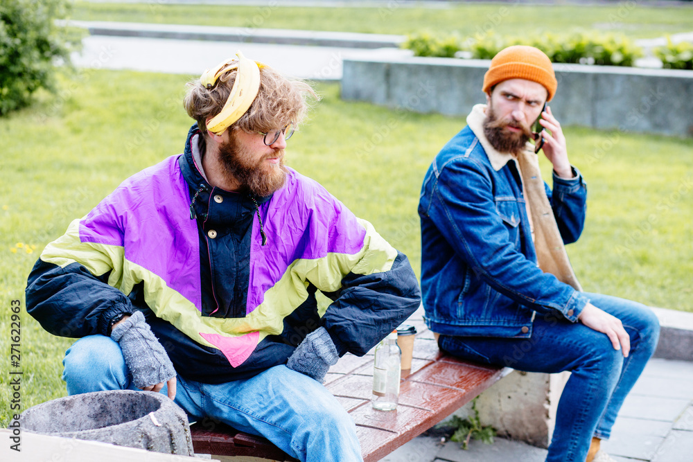 Homeless man with banana skin near full garbage basket in colorful clothes rummaging in trash container in park green background. Shocked hansome beard male sitting on the bench near him