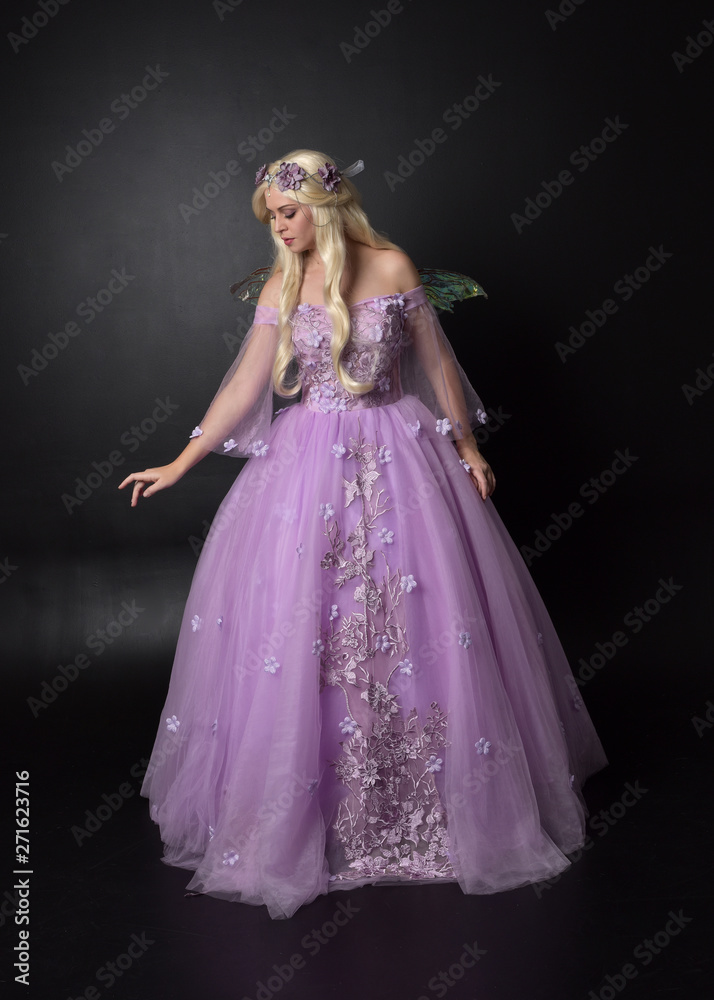 full length portrait of a blonde girl wearing a fantasy fairy inspired costume, long purple ball gown with fairy wings, standing pose on a dark studio background. full length portrait of a blonde girl wearing a fantasy fairy inspired costume, long purple ball gown with fairy wings, standing pose on a dark studio background.