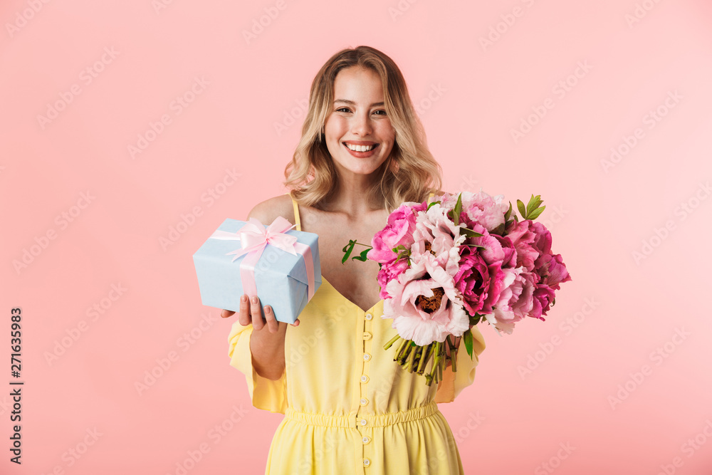 Young blonde woman posing isolated over pink wall background holding flowers holding present gift box. Young blonde woman posing isolated over pink wall background holding flowers holding present gift box.