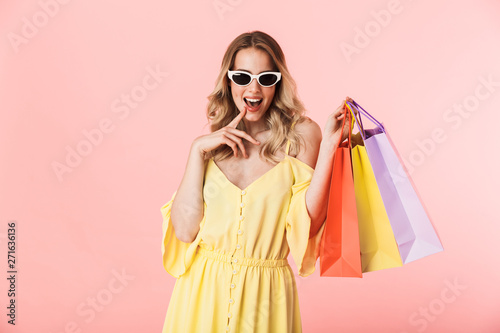 Beautiful happy young blonde woman posing isolated over pink wall background holding shopping bags.