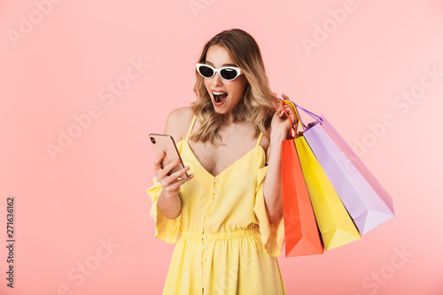 Excited happy young blonde woman posing isolated over pink wall background holding shopping bags using mobile phone.