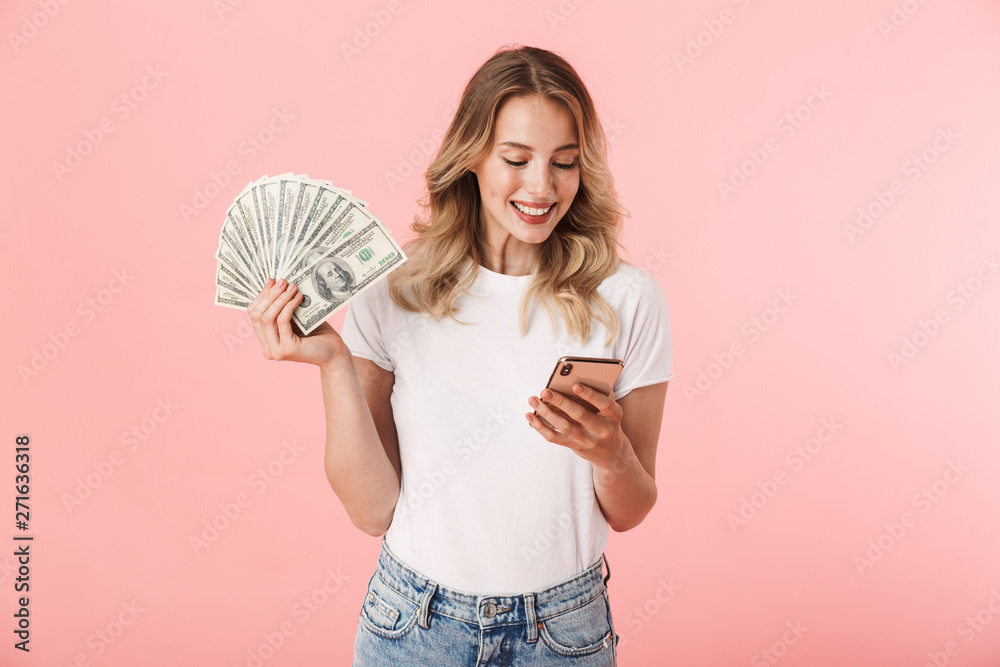 Excited young blonde woman posing isolated over pink wall background holding money and using mobile phone. Excited young blonde woman posing isolated over pink wall background holding money and using mobile phone.
