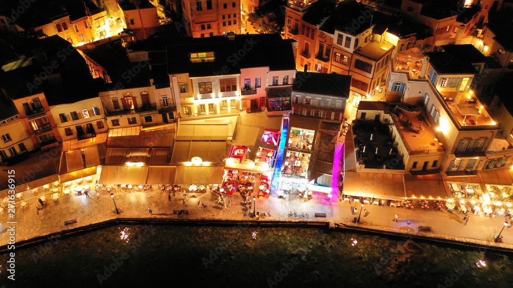 Obraz premium Aerial drone night shot of iconic and picturesque illuminated Venetian old port of Chania with famous landmark lighthouse and shops - restaurants serving Cretan traditional food, Crete island, Greece