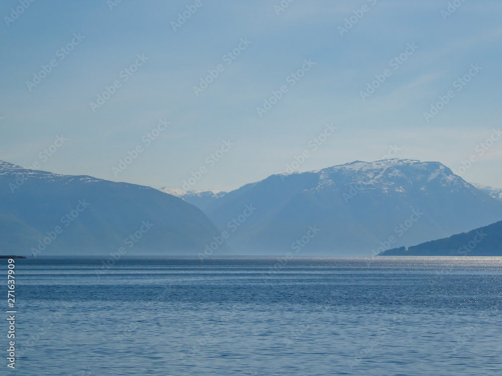 A view on the Songefjorden (King of the Fjords) from the water level. It is the deepest fjord in Norway. Tall, lush green mountains surrounding the fjord. Calm surface of the water. Clear blue sky.