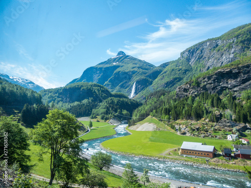 A little village located at the bottom of a valley, with a river flowing in the middle. Tall mountains surrounding the village. Clear and bright day.