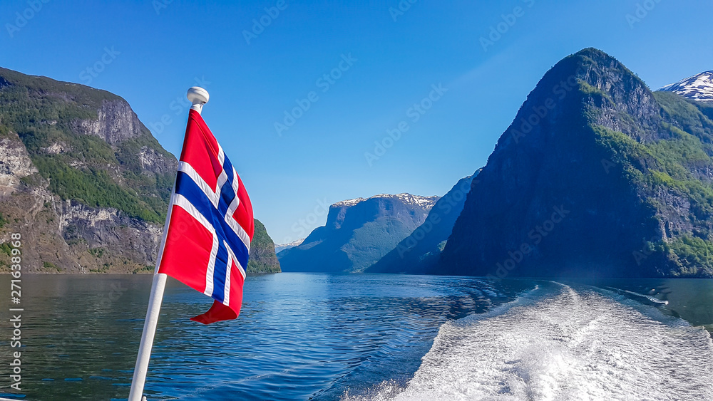Norwegian flag hanging on  the railing of the ship and waving above the water.The motor of the ship makes the water wavy and foamy. Tall, lush green mountains surrounding the fjord. Clear blue sky.