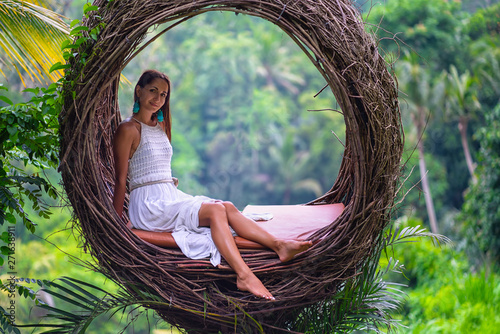 Beautiful tan woman in white dress posing on a straw nest. Rain forest on the background