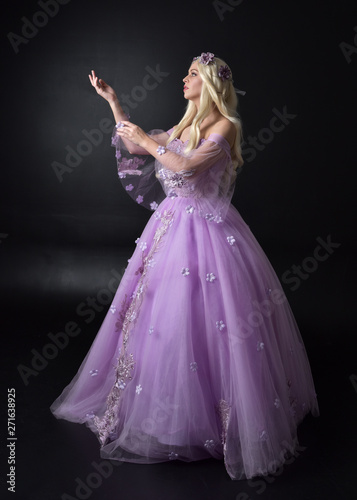 full length portrait of a blonde girl wearing a fantasy fairy inspired costume, long purple ball gown with fairy wings, standing pose on a dark studio background.