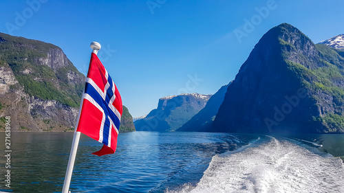 Norwegian flag hanging on  the railing of the ship and waving above the water.The motor of the ship makes the water wavy and foamy. Tall, lush green mountains surrounding the fjord. Clear blue sky. photo