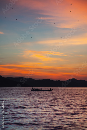 Landscape with a boat floating on the water. On the background of the contour of the shoreline and a fiery sunset. Vertical orientation