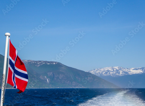 Norwegian flag hanging on the railing of the ship and waving above the water.The motor of the ship makes the water wavy and foamy. Tall, lush green mountains surrounding the fjord. Clear blue sky.