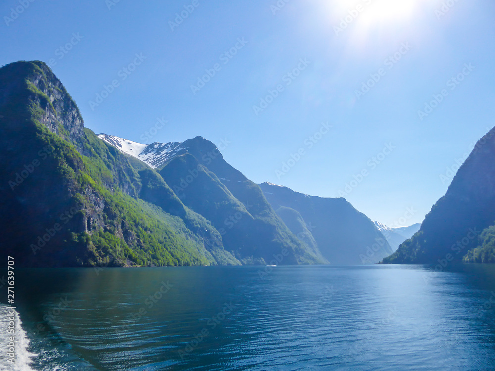 A view on the Songefjorden fjord from the water level. The motor of the ship makes the water wavy and foamy. Tall, lush green mountains surrounding the fjord. Clear blue sky.