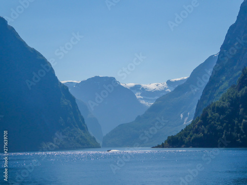 A view on the Songefjorden (King of the Fjords) from the water level. It is the deepest fjord in Norway. Tall, lush green mountains surrounding the fjord. Calm surface of the water. Clear blue sky.
