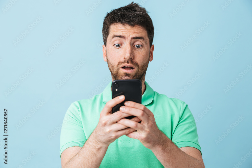 Shocked scared young handsome bearded man posing isolated over blue wall background using mobile phone. Shocked scared young handsome bearded man posing isolated over blue wall background using mobile phone.