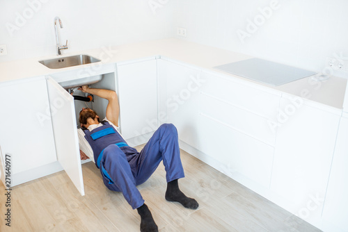 Plumber in overalls repairing or installing sewerage under the kitchen sink at home