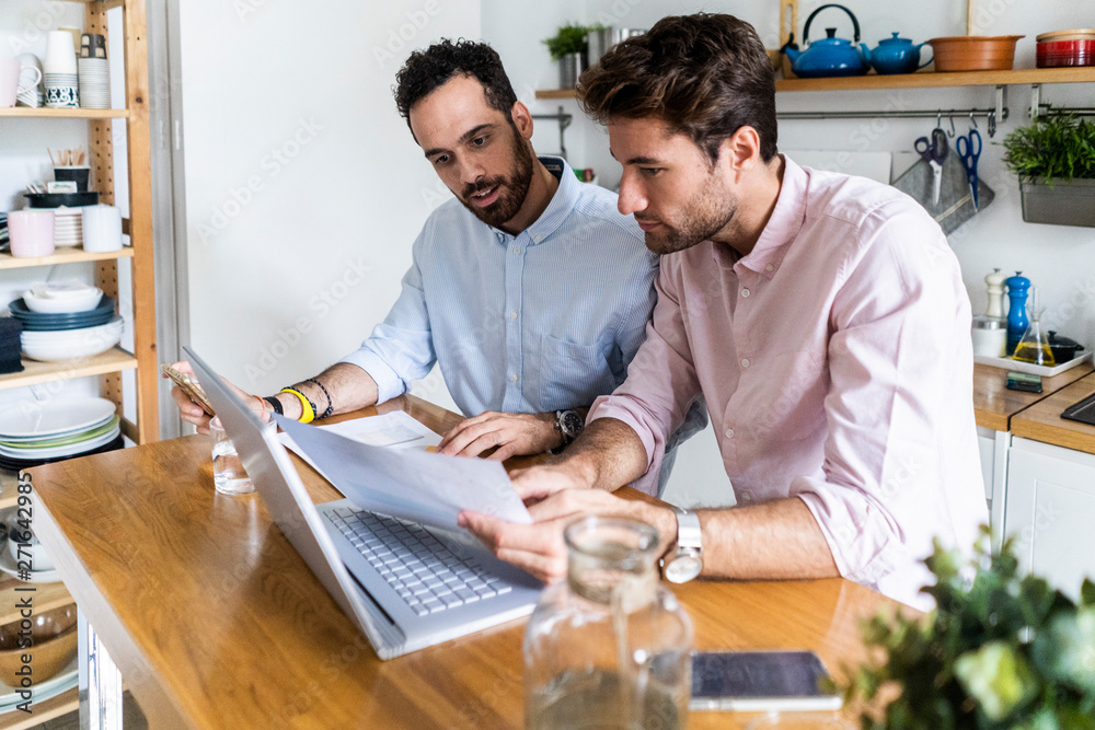 Two young men working together in kitchen, using laptop, discussing documents Two young men working together in kitchen, using laptop, discussing documents