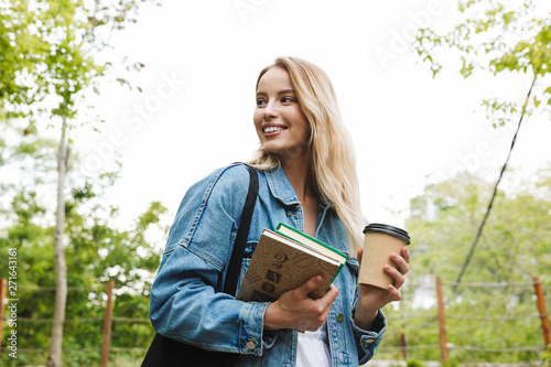 Canvas Print Happy young woman student posing outdoors in park drinking coffee holding books