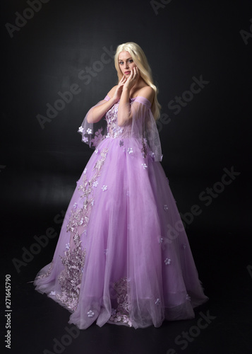 full length portrait of a blonde girl wearing a fantasy fairy inspired costume, long purple ball gown with fairy wings, standing pose on a dark studio background.