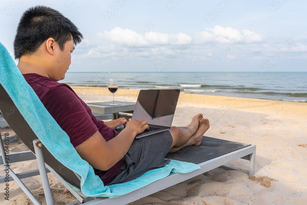 Asian man using laptop relaxing on bench by beautiful beach