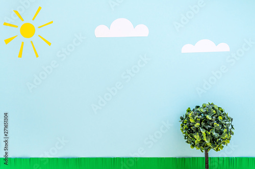 Paper sun, clouds, green grass and toy tree with green leaves on a blue background. Funny summer landscape composition on the theme of good sunny weather and good mood.