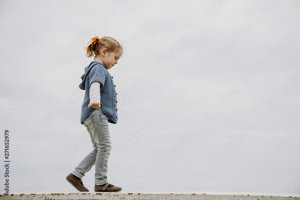 Side view of little girl with outstretched arms walking on border against calm sea and gray sky Side view of little girl with outstretched arms walking on border against calm sea and gray sky