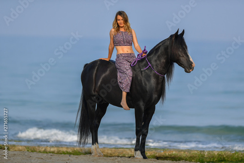 Portrait of a beautiful blond girl sitting on the back of her black horse on the sea beach