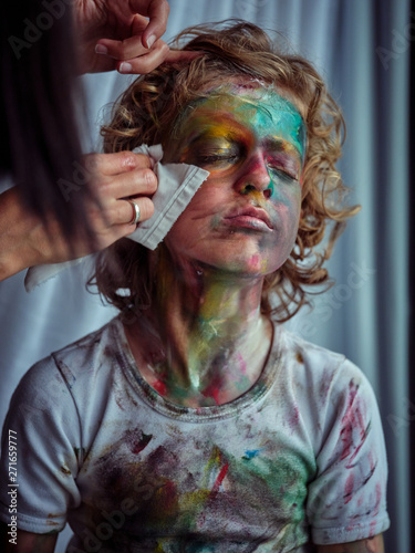 Crop young beautiful female with napkins cleaning pretty creative boy with blond hair smeared in colorful paints photo