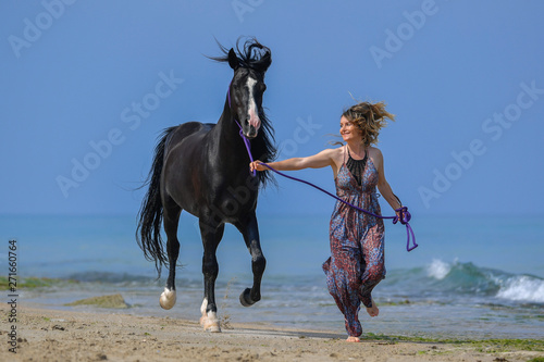 Portrait of a beautiful blond girl running on the sand with her black horse on the sea beach
