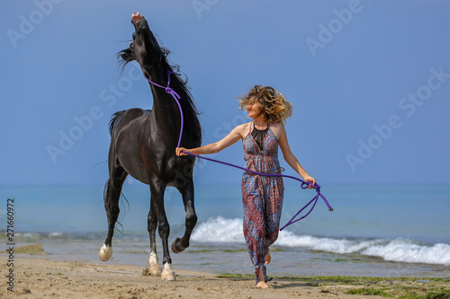 Portrait of a beautiful blond girl running on the sand with her black horse on the sea beach