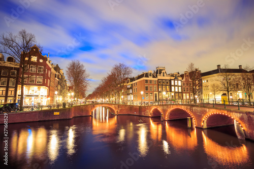 Illuminated bridges on intersecting canals, under a dramatic sky at night, in Amsterdam, Netherlands.