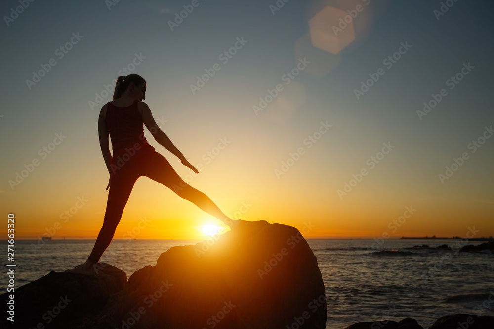 Silhouette of young woman doing fitness exercises on the beach at amazing sunset. Yoga and health.