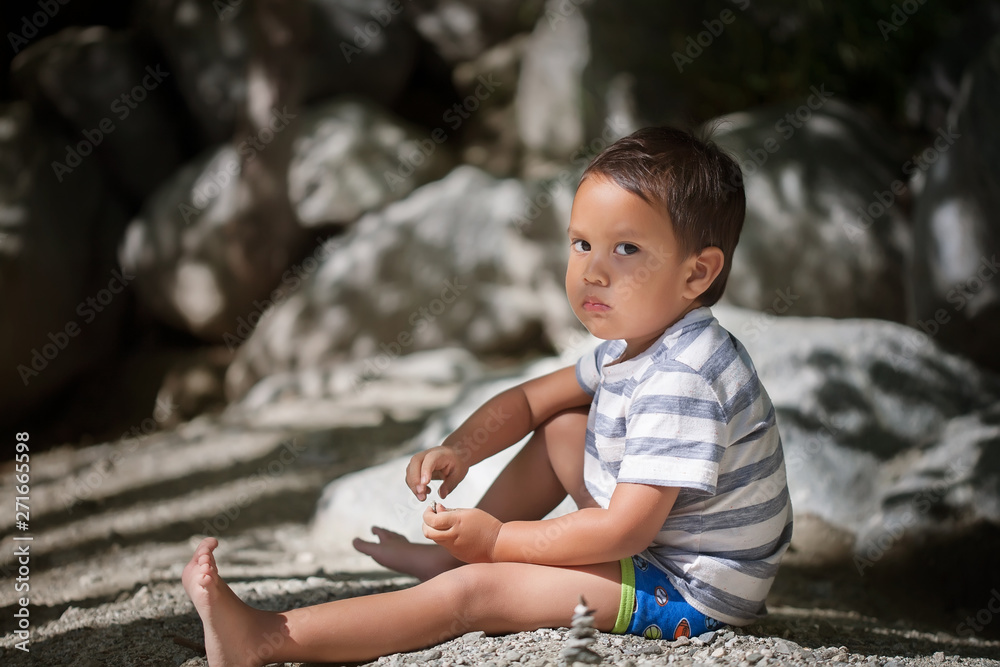 An upset little boy with a moody expression on his face, wearing a T-shirt and shorts while sitting alone on the shore next to big rocks. An upset little boy with a moody expression on his face, wearing a T-shirt and shorts while sitting alone on the shore next to big rocks.
