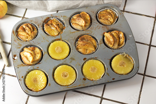 Gluten-free homemade vegetarian desserts cupcakes muffins with apple slices and pineapple rings on top in baking tray in kitchen on white-tiled worktop, next to a kitchen tea-strainer, towel