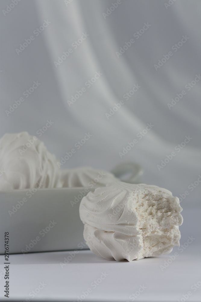 Obraz premium One bitten marshmallow with full square deep plate of marshmallows on white background. Natural daylight.