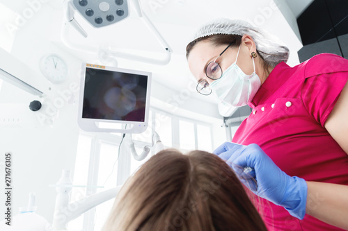 A professional dentist woman in glasses and overalls examines the oral cavity of a young girl in the dental chair using an intraoral stamotological video camera with LED illumination