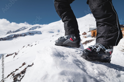 A close-up of a skier's leg in ski boots without skis stands on a knoll of bliss against the background of mountains on a sunny day. The concept of skittura and hiking ski travel