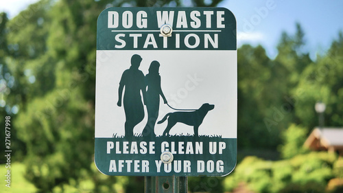A green and white sign that says Dog Waste Station, please clean up after your dog. Background is green treed neighborhood.