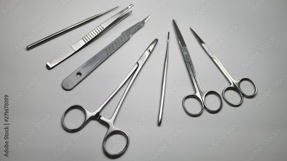 A pair of stainless steel surgical clamps, scalpel knife, medical grade scissors, forceps and assorted probes on a white table. A pair of stainless steel surgical clamps, scalpel knife, medical grade scissors, forceps and assorted probes on a white table.