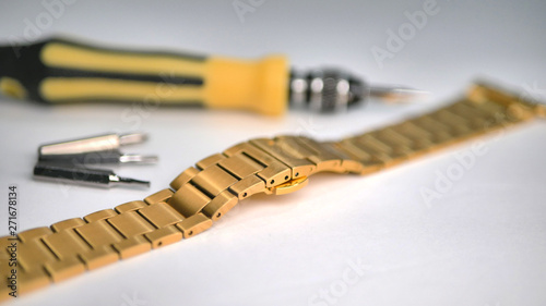 A shiny gold colored metal chain link wrist watch band with repair screwdriver laying on a white table.