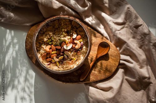 Oatmeal porridge in coconut bowl with wooden spoon on natural wooden tray. Porridge oats with almonds, pistachios and other nuts. Shadow of sunrise morning. White background. Top view or flat lay.