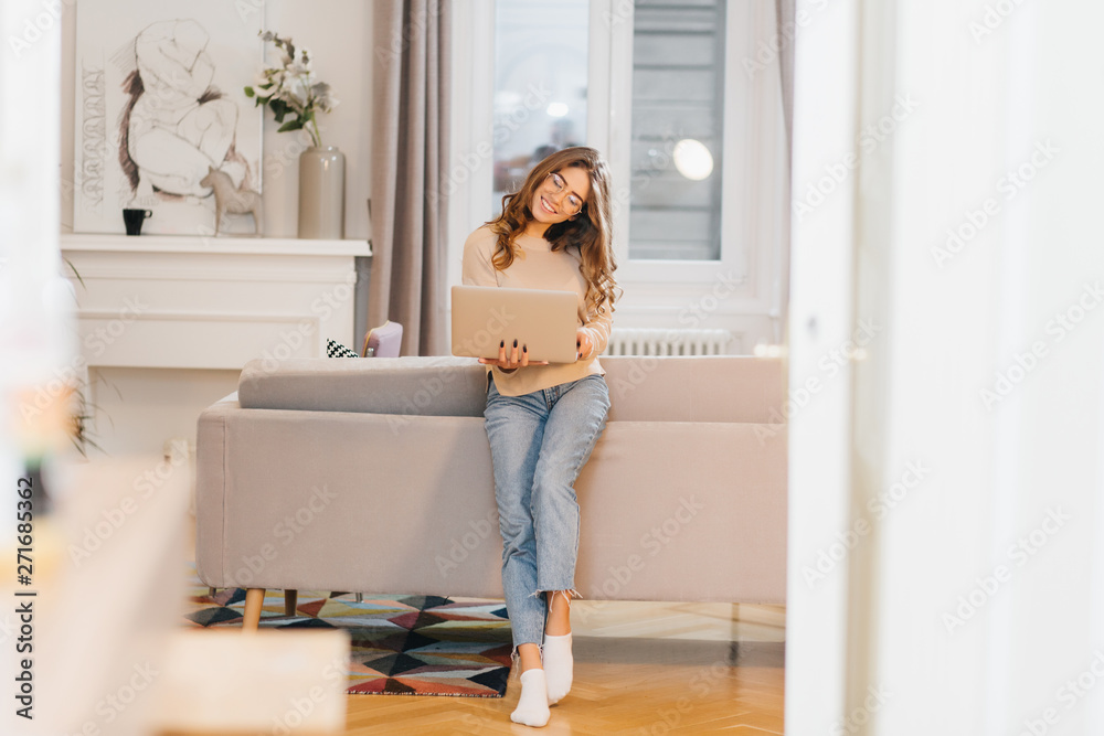 Romantic female student in white socks holding laptop and smiling. Full-length portrait of trendy brunette girl in jeans using computer in living room. Romantic female student in white socks holding laptop and smiling. Full-length portrait of trendy brunette girl in jeans using computer in living room.