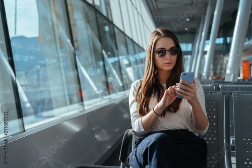 Young passenger woman in casual clothes sitting at the gate and using smartphone while waiting for a flight at the airport. Travel concept. Girl chatting in terminal departure lounge.