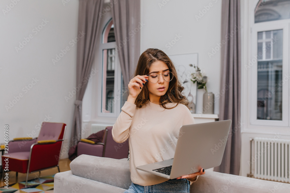 Serious smart woman in glasses posing with laptop. Indoor portrait of elegant female model looking at computer screen while standing in cozy room.