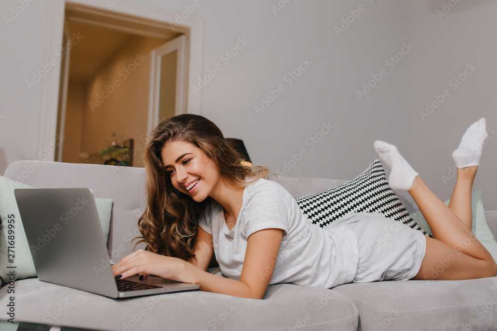 Lovely brunette girl in white socks using laptop with charming smile. Pleased young lady posing on gray couch with cushions and typing on keyboard. Lovely brunette girl in white socks using laptop with charming smile. Pleased young lady posing on gray couch with cushions and typing on keyboard.