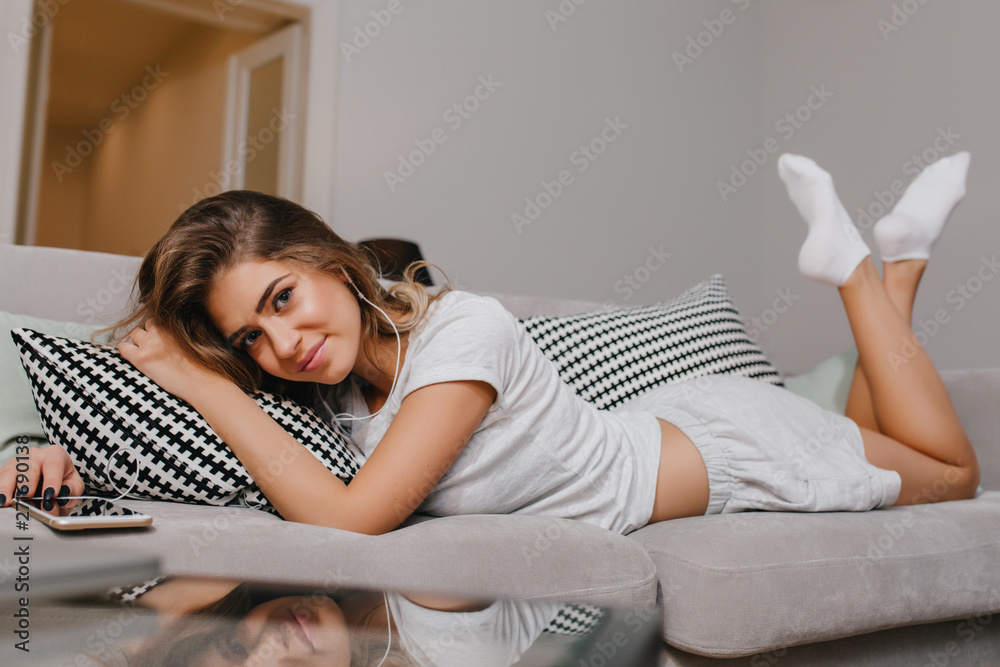 Good-looking young woman in white pajamas lying on sofa and listening favorite song. Photo of pretty caucasian female lady in socks resting after hard day on couch. Good-looking young woman in white pajamas lying on sofa and listening favorite song. Photo of pretty caucasian female lady in socks resting after hard day on couch.