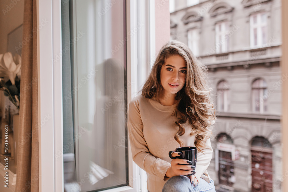 Interested shy girl with long hair posing with cup of tea on sill. Indoor portrait of good-looking curly model in beige shirt sitting near window on city background.
