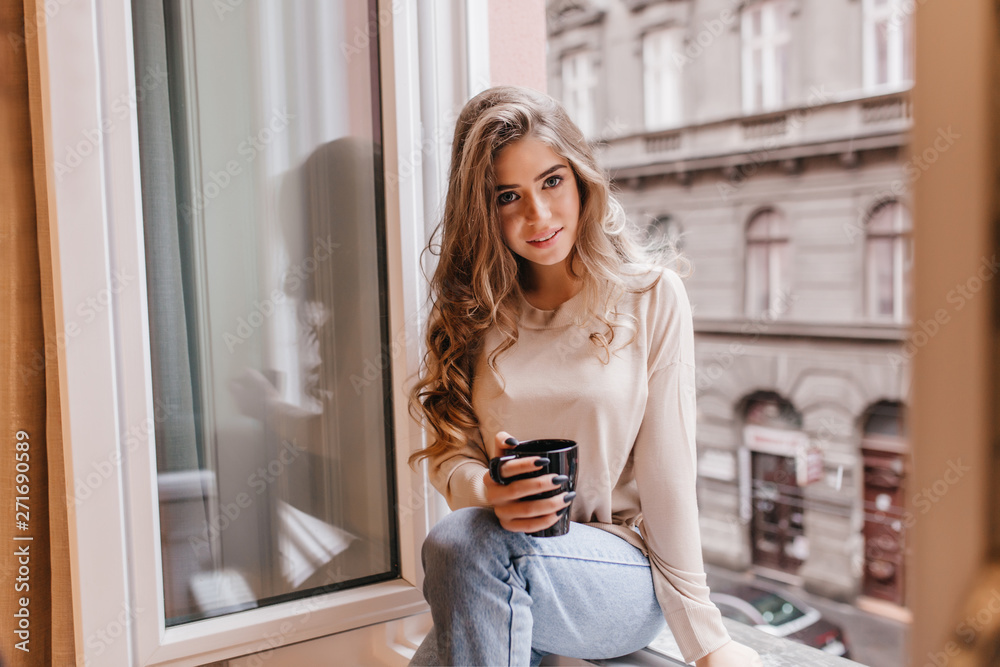 Shy caucasian girl in jeans sitting on window sill and posing with interest. Indoor portrait of trendy white woman with cup of cappuccino relaxing in morning. Shy caucasian girl in jeans sitting on window sill and posing with interest. Indoor portrait of trendy white woman with cup of cappuccino relaxing in morning.