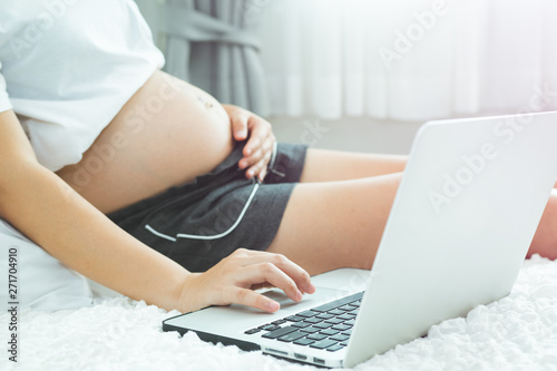 portrait of an Asian pregnant woman using laptop on the bed surfing internet and take hands rub her stomach